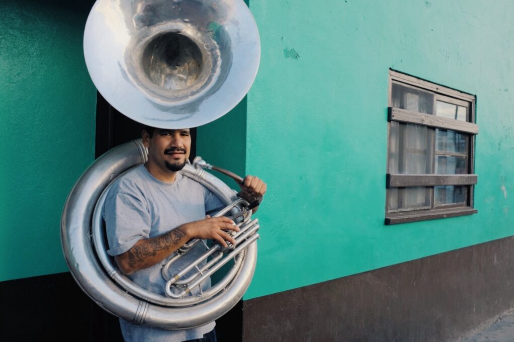 Man with sousaphone stands against a teal wall