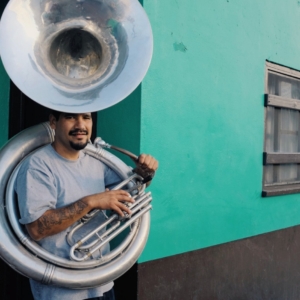 Man with sousaphone stands against a teal wall