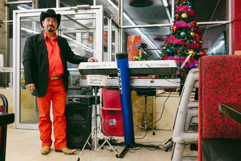 Man in red shirt and pants and black blazer and cowboy hat stands in front of a Casio keyboard